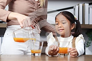 Asian happy family spending time together, mother pouring fresh orange juice from jug for her little cute daughter sitting in