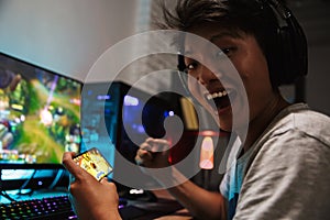 Asian happy gamer boy rejoicing while playing video games on smartphone and computer in dark room, wearing headphones and using b