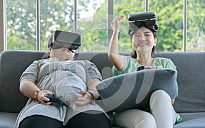 Asian happy lovely mother and young chubby down syndrome autistic autism little daughter sitting on cozy sofa wearing VR virtual