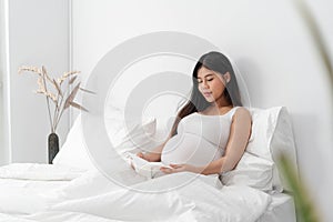 Asian happy pregnant woman is sitting and reading a book on bed and and touching her belly