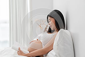 Asian happy pregnant woman is sitting and reading a book on bed and and touching her belly. pregnancy