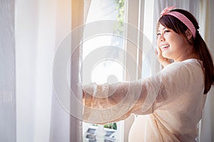 Asian happy pregnant woman standing beside window and open curtain in the morning