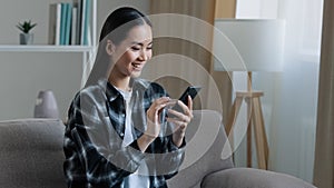 Asian happy smiling girl woman sitting home sofa looking at mobile phone chatting browsing using wifi web service