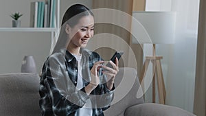 Asian happy smiling girl woman sitting home sofa looking at mobile phone chatting browsing using wifi web service