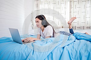 Asian happy young woman wear headphones and listening music song and watching website for shopping online from laptop  on bed at