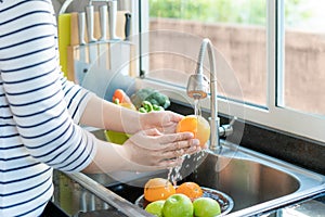 Asian healthy woman washing an orange and other fruit above kitchen sink and cleaning a fruit / vegetable with water to eliminate