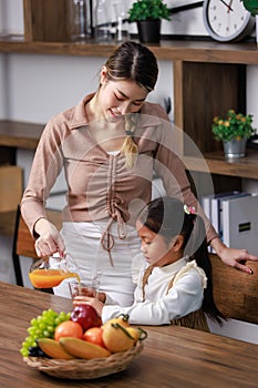 Asian housewife standing smiling near bookshelf pouring organic orange juice to little girl kid daughter in living room at home
