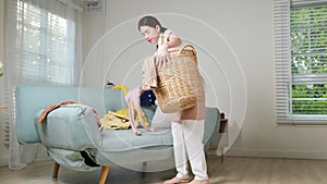 Asian housewife woman holding basket with heap of different clothes on sofa in living room