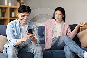 Asian Husband Texting On Smartphone Ignoring His Displeased Wife Indoor