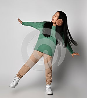 Asian kid girl in green sportswear and white sneakers stands on one leg with arms outstretched, balancing