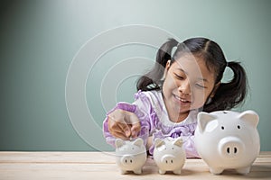 An Asian kid girl put a coin in a white pig piggy bank on a wood table at home. Save money for tuition fees, the cost of school