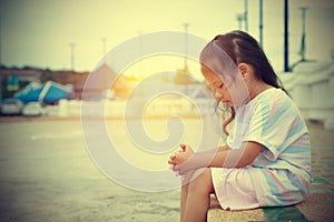 An Asian kid girl is sitting alone on a sidewalk. A sad face unhappy.