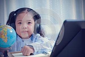 Asian kid girl wearing headphones looking at the camera learning education distance from home by tablet. Current kids around the