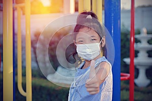 Asian kid girl wearing a medical mask protect coronavirus, sitting alone, looking at the camera in the garden show thumbs up for