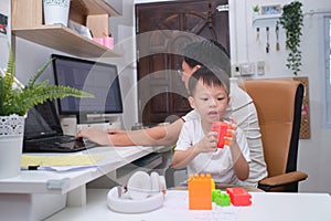 Asian kindergarten boy having fun playing blocks near his working at home father, while in quarantine isolation during the Covid-