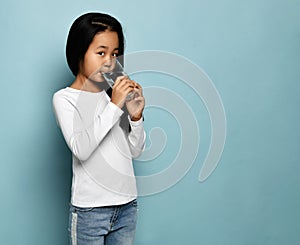 Asian Korean kid girl in jeans and white long sleeve shirt stands drinking pure water from glass at copy space