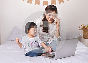Asian lady in classic suit is talking on the mobile phone and working on laptop at home with her baby girl.