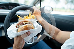 Asian lady holding hamburger and French fries to eat in car, dangerous and risk an accident