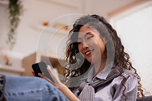 Asian lady sits on sofa at home, texting and talking with buddy, holding smartphone, shopping online, or checking mobile app or