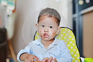 Asian little baby boy sitting on children chair indoor eating bread with Stuffed Chocolate-filled dessert and Stained around her