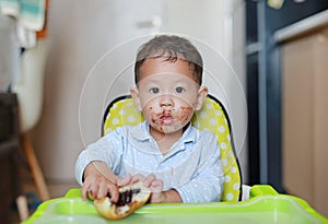 Asian little baby boy sitting on children chair indoor eating bread with Stuffed Chocolate-filled dessert and Stained around her