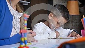 Asian little boy writing homework while mother explaining something to him, sitting behind table, book shelves and brick