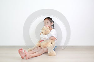 Asian little child girl hugging a teddy bear doll sitting against white wall in the room