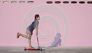 Asian little dark skinned girl has fun by riding kick scooter near pink wall on the patio