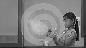 Asian little dark-skinned girl using spray bottle spraying water into her cactus plant inside of glass window frame at home in