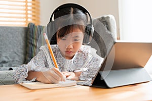 Asian little girl doing homework, using digital tablet with headphone. Homeschooling, Concept online learning at home