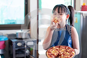 Asian little girl eating pizza with delicious and happiness after making homemade pizza in the home kitchen