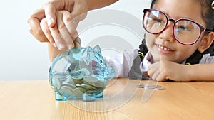 Asian little girl in Thai kindergarten student uniform putting money coin into clear piggy bank on wooden table metaphor money sav