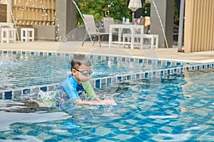 Asian little kid in swimming goggles and swimming suit learning to swim at outdoor pool in summer