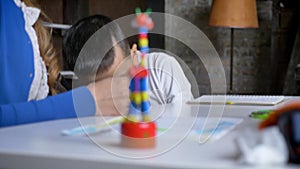 Asian little kid taking falled pen from floor and studing with mother, sitting behind table, book shelves and brick wall