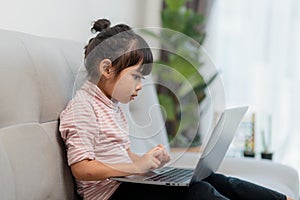 Asian Little school kid girl use laptop computer sitting on sofa alone at home. Child learning reading online social media content