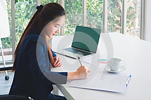 Asian long hair beautiful business woman in navy blue suit working, writing document on the table in office. Tree outside windows.