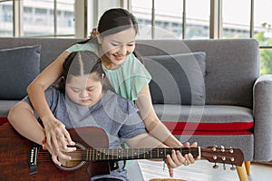 Asian lovely mother helping young chubby down syndrome autistic autism little daughter holding playing guitar in living room at