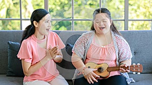 Asian lovely mother holding guitar sit smiling on sofa couch in living room at home teaching young chubby down syndrome autistic