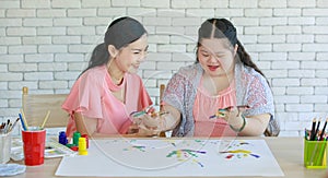 Asian lovely ponytail hair mother using paintbrush painting acrylic colors on young cute chubby down syndrome autistic daughter