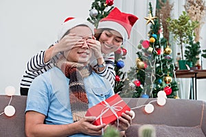 Asian lovers couple, girlfriend surprise boyfriend by giving Christmas present at sofa with xmas decoration tree at house