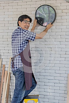 Asian male adult wearing clear safety glasses and blue plaid shirt holding and adjusting black round metal clock with white