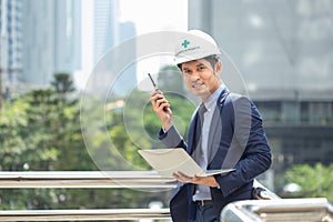 Asian male architect in suit wearing helmet standing holding a radio and laptop, portrait man looking camera.