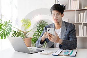 Asian male businessman using smartphone to do financial work on laptop