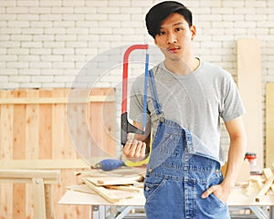 Asian male carpenter wearing denim bib pants standing in wood workshop holding hand saw and looking at camera