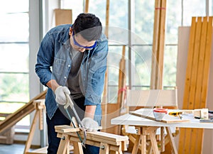 Asian male carpenter woodworker engineer in jeans outfit with safety gloves and glasses goggles using hand saw cutting wood stick