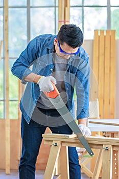 Asian male carpenter woodworker engineer in jeans outfit with safety gloves and glasses goggles using hand saw cutting wood stick