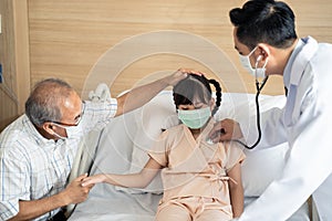 Asian male doctor with mask coming to recovery room and asking health problem to young kid patient on bed with parent in hospital