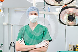 Asian male doctor standing with his arms crossed wearing a green surgical gown in the operating room