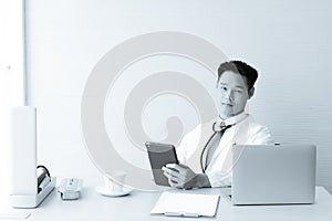 Asian male doctor with stethoscope looking at camera sat on the working chair hold a tablet on a desk in the hospital,monochrome
