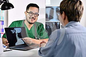 Asian male doctor in surgical uniform consulting and explaining female patient using laptop in examination room